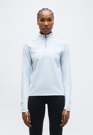 Femeie cu părul împletit, purtând un top cu mâneci lungi, bleu deschis, cu fermoar și pantaloni negri, stând cu fața la cameră pe un fundal simplu.