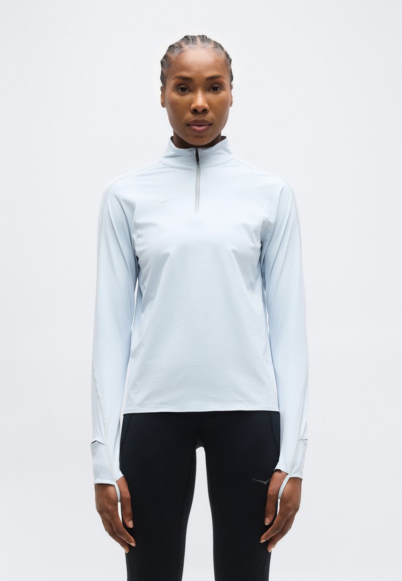 Γυναίκα με πλεξούδες που φοράει ανοιχτό γαλάζιο μακρυμάνικο μπλουζάκι με φερμουάρ και μαύρο παντελόνι, όρθια, κοιτάζοντας προς τα εμπρός, μπροστά σε ένα απλό φόντο.