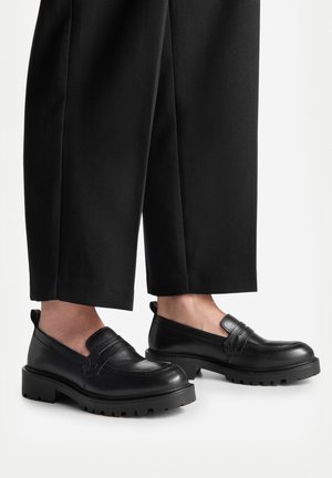 Stopy w czarnych skórzanych lordach z masywnymi gumowymi podeszwami, zestawione z czarnymi spodniami o szerokich nogawkach i skróconej długości, na jednolitym tle.