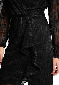 Robe noire en tissu texturé et plissé. Dotée d&#x27;un détail volanté le long de l&#x27;ourlet et de manches longues transparentes avec une taille ajustée.