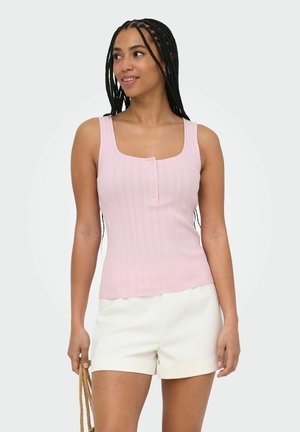 Femme aux cheveux tressés portant un haut sans manches rose clair boutonné et un short blanc, tenant la poignée d’un sac tressé.