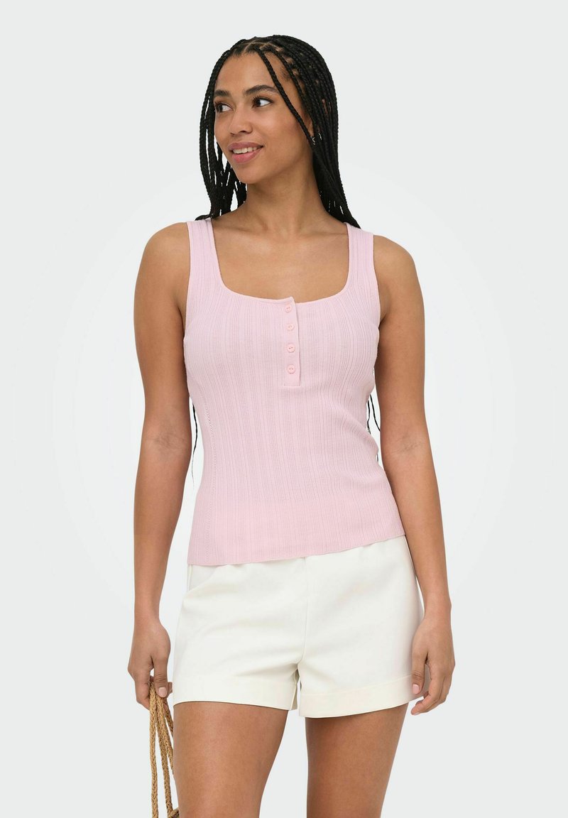 Femme aux cheveux tressés portant un haut sans manches rose clair boutonné et un short blanc, tenant la poignée d’un sac tressé.
