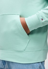 Άτομο που φοράει ανοιχτό πράσινο φούτερ με το χέρι στην μπροστινή τσέπη και μικρό κόκκινο, λευκό, μπλε λογότυπο στην μανσέτα του μανικιού, συνδυασμένο με μπλε τζιν.