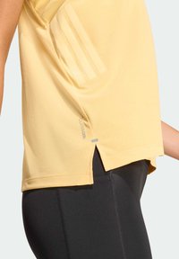 Camiseta deportiva de color amarillo claro con mangas cortas, tejido texturizado, abertura lateral en el dobladillo y tres rayas tonales en el lado izquierdo.