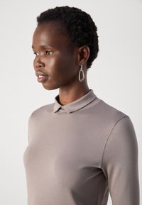 Femme aux cheveux courts et bouclés portant des boucles d'oreilles en argent et un haut ajusté à manches longues taupe avec un petit col, regardant vers la gauche.