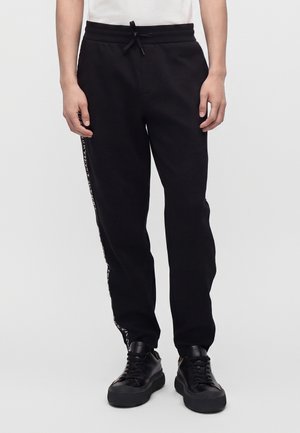 Sorte joggerbukser med sidestrejfdetalje, båret med sorte sneakers og en hvid top, vist fra talje til fødder.