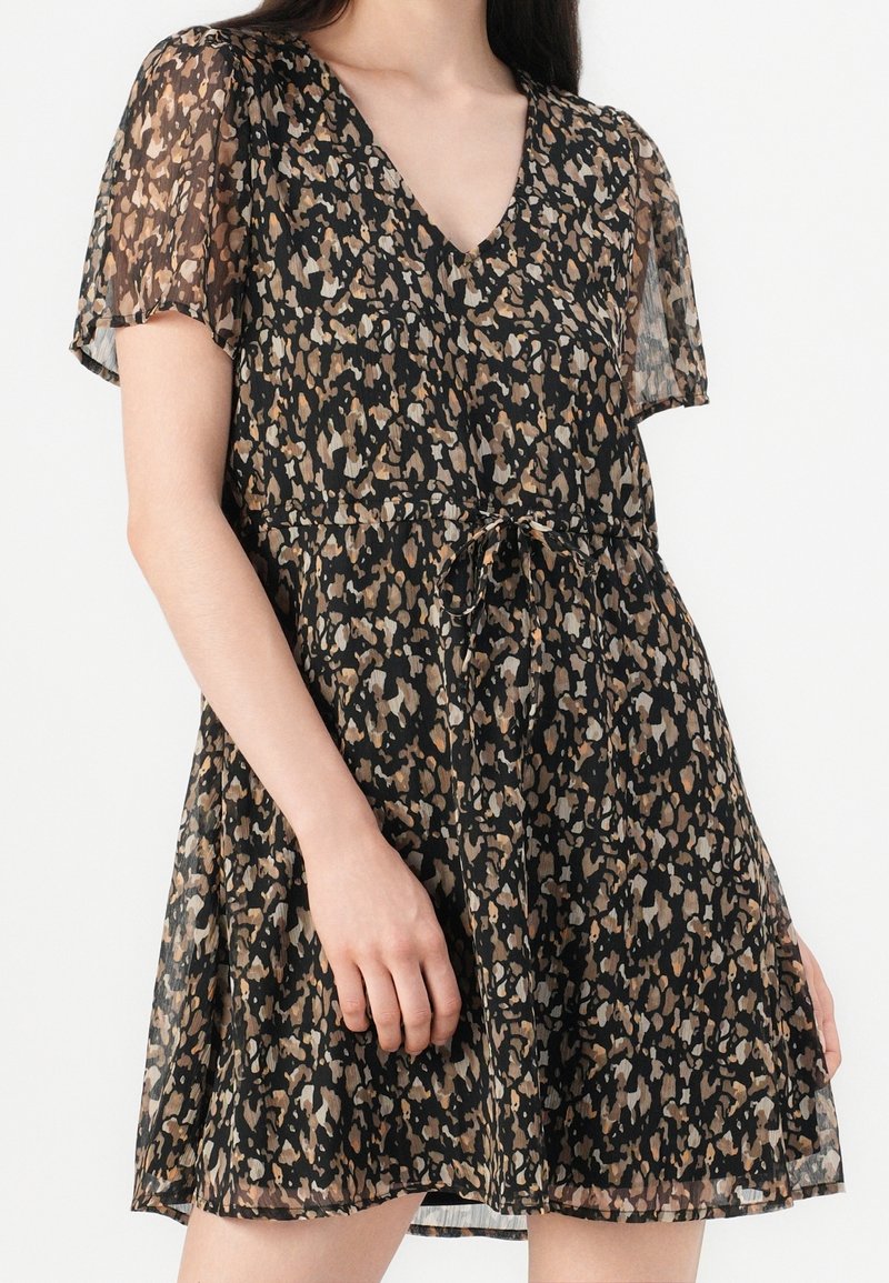 Femme portant une robe noire à manches courtes et col en V avec des motifs abstraits marron et beige et une ceinture nouée à la taille.