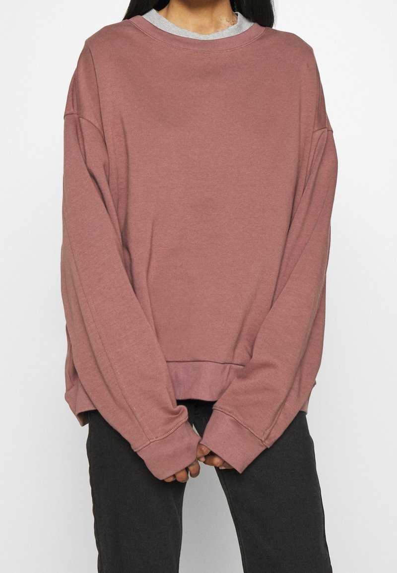 Personne portant un sweat-shirt oversize rose fané avec des épaules tombantes et un pantalon foncé, les mains jointes devant à la taille.