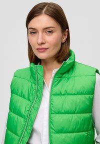 Grüne plüschige Weste mit hohem Kragen, Reißverschluss und horizontalem Steppmuster. Getragen über einem weißen Hemd mit glänzender, glatter Textur.