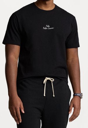 Mężczyzna w czarnej koszulce Polo Ralph Lauren z białym logo i czarnych spodniach z białym sznurkiem, ręce luźno opuszczone wzdłuż ciała.