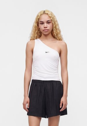 Mladá žena s dlhými kučeravými blond vlasmi, ktorá má na sebe biely top Nike na jedno rameno a čierne šortky, stojí na jednoduchom pozadí.