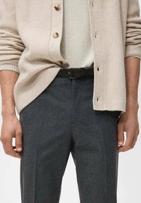 Beżowy kardigan na guziki nałożony na dopasowany, dziany sweter, zestawiony z ciemnoszarymi, dopasowanymi spodniami i czarnym paskiem, z rękami opuszczonymi wzdłuż ciała.