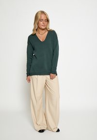 Dunkelgrüner V-Ausschnitt-Pullover aus weichem Stoff, kombiniert mit lockeren beigen Hosen und schwarzen Schuhen, die eine entspannte Outfit-Silhouette kreieren.