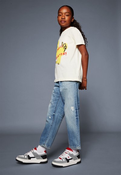T-shirt color crema con grafica gialla e testo rosso, abbinata a jeans distressed blu chiaro e sneakers grigio-nero-rosse.