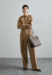 Chemise beige cintrée et pantalon large taille haute. Sac fourre-tout texturé marron avec des anses en cuir. Fond de couleur unie.