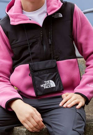 Personne portant une veste polaire rose et noire et un pantalon gris, avec une petite pochette noire étiquetée "The North Face" suspendue au cou.