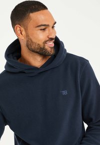 Marine fleece hoodie met een ruime capuchon, textuurstof en een geborduurd TB-logo op de borst. Zacht en casual ontwerp.