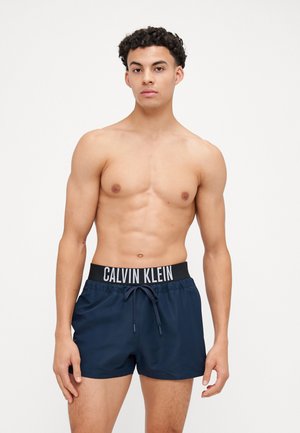 Marineblå badebukser med en sort elastik, der har den hvide tekst "CALVIN KLEIN." Designet inkluderer en snørelukning til justering.