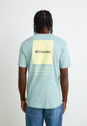 T-shirt vert menthe à manches courtes avec un grand rectangle de logo jaune pastel au dos, contenant "Columbia" et du texte en dessous en caractères blancs.
