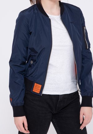 Marineblauwe bomberjack met ritssluiting, zijzakken, zwarte geribbelde manchetten en een oranje labeldetail aan de linkerkant. Glad, glanzend oppervlak.