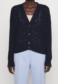 Gilet en tricot bleu marine foncé avec fermeture à boutons sur un haut beige, associé à un pantalon light blue ajusté, porté par une personne de profil.
