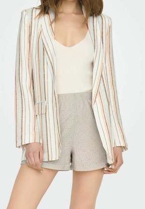 Vrouw die een gestreepte blazer met pastelkleurige tinten draagt, een witte geribbelde tanktop en beige shorts van eyelet-stof, staand tegen een witte achtergrond.