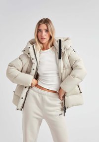 Puffet beige jakke med hætte og sorte detaljer, med frontlynlås og trykknapper, parret med bløde, lysegrå sweatpants.