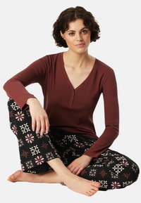 Bordeaux langwerpige top met knopen bij de halslijn, gecombineerd met zwarte broek met kleurrijke bloemenpatronen. Model zit.