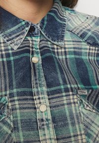 Chemise à carreaux aux tons bleu, vert et blanc ; présente un devant à boutons, un col classique et une étiquette de marque sur le côté. Tissu texturé.