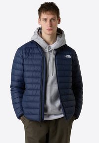 Giacca a vento blu navy con design trapuntato, chiusura con zip, colletto alto e logo The North Face. Indossata sopra un hoodie grigio.