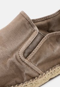 Zapato slip-on de lona marrón claro con un exterior texturizado, elasticidad lateral y suela de cuerda de yute. Detalle de costura sutil a lo largo de los bordes.
