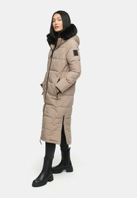 Langer beige Steppmantel mit schwarzem Kunstfellkragen, Reißverschluss, zwei Seitentaschen und gerippten Akzenten. Ein Paar schwarze Knöchelstiefel sind sichtbar.