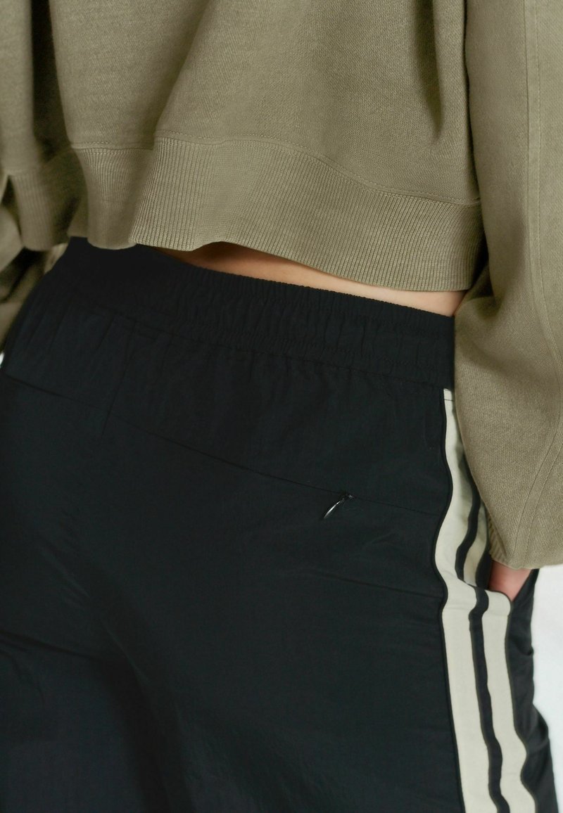 Personne portant un pantalon noir avec des bandes latérales beige et noire et un pull court vert olive, main dans la poche.