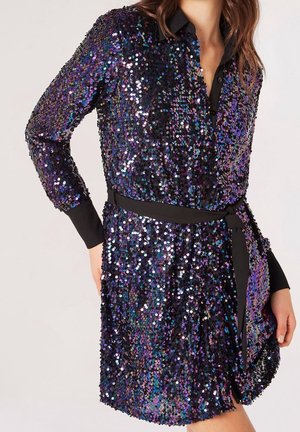 Femme portant une robe noire à manches longues en sequins aux nuances irisées violet et bleu avec une ceinture noire à la taille, debout avec une main sur la hanche.
