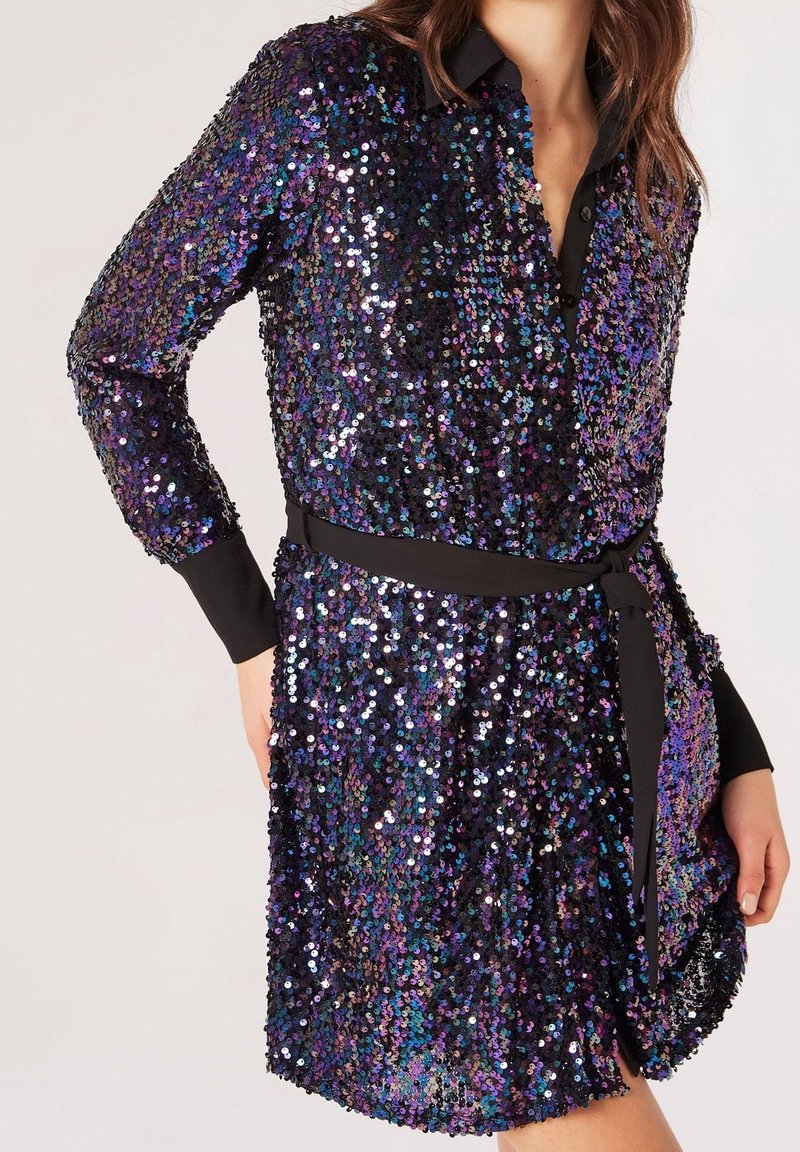 Femme portant une robe noire à manches longues en sequins aux nuances irisées violet et bleu avec une ceinture noire à la taille, debout avec une main sur la hanche.