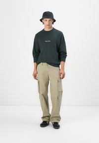 Sudadera verde oscura con cuello redondo y texto bordado en blanco, combinada con pantalones cargo beige que tienen múltiples bolsillos. Se llevan zapatillas negras.