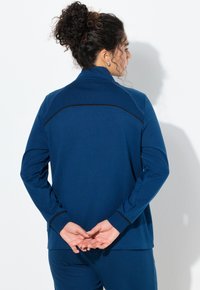 Chaqueta azul con cremallera y mangas largas, hecha de tela lisa. Cuenta con una franja negra contrastante en la parte superior de la espalda.