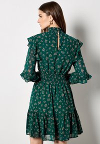 Zielona sukienka w kwiaty z falbanowymi rękawami, marszczonym pasem i warstwową spódnicą. Detal w kształcie łezki z tyłu oraz drobne wzory kwiatowe na całej powierzchni.