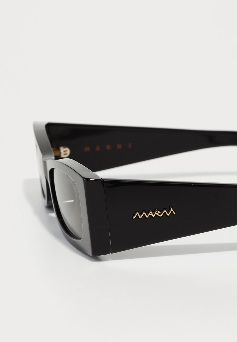 Lunettes de soleil noires rectangulaires avec des montures en plastique brillant, des accents argentés et un logo doré visible sur la branche. Les verres sont légèrement teintés.