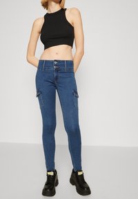 Jeans de tiro alto azul con un ajuste ajustado, que presentan bolsillos tipo cargo y cierre de botón. Combinados con una camiseta sin mangas negra y botas negras gruesas.