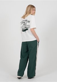 Hvit T-skjorte med korte ermer og svart grafisk trykk på baksiden, kombinert med løse grønne bukser med detaljert sidestripe.