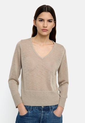 Młoda kobieta o długich ciemnych włosach, ubrana w beżowy sweter z dekoltem w serek i niebieskie dżinsy, stoi z rękami w kieszeniach na białym tle.