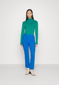 Pull à col roulé vert associé à un pantalon bleu sur mesure, avec une coupe ajustée et une texture lisse, complété par des bottes à talons beiges.