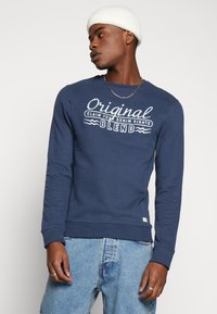 Tamno plavi dukserica od teksturirane pamučne tkanine, sa bijelim otisnutim tekstom "Original Claim Your Denim Rights" preko prsa i rebrastim rukavima.