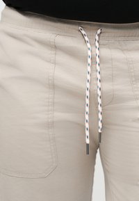 Gros plan sur un pantalon beige avec un cordon de serrage blanc orné de motifs bleus et rouges, montrant les poches avant et les détails de couture.