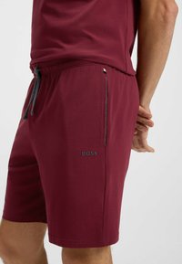 Burgundy shorts i mjukt tyg med elastisk midja, grå dragband och en 'BOSS' logotyp på vänster lår. Slät yta.