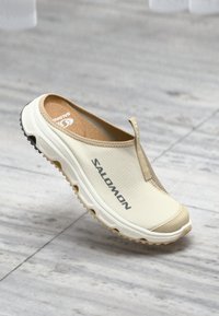 Béžová obuv na slip-on s textúrovaným zvrškom, gumovou podrážkou a viditeľným logom. Obsahuje slučku pre jednoduché obúvanie a vypchávku vo vnútri.