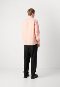 Ung mann som står med ryggen til i en lys rosa skjorte med lange ermer, sort bukser og svarte sko mot en enkel hvit bakgrunn.
