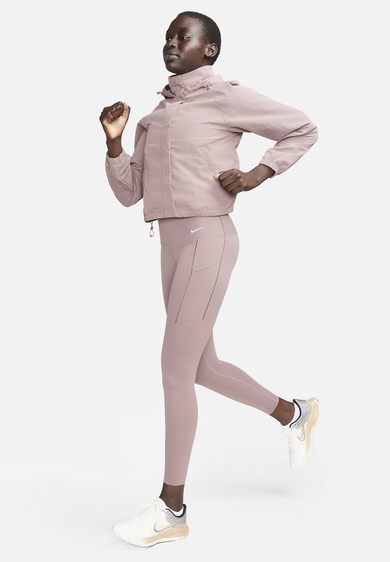 Vrouw die jogt in een mauve jack en legging met witte en beige hardloopschoenen tegen een effen achtergrond.