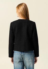 Schwarze strukturierte Jacke mit langen Ärmeln, geradem Schnitt und rundem Ausschnitt. Über blauen Jeans getragen. Rückansicht.